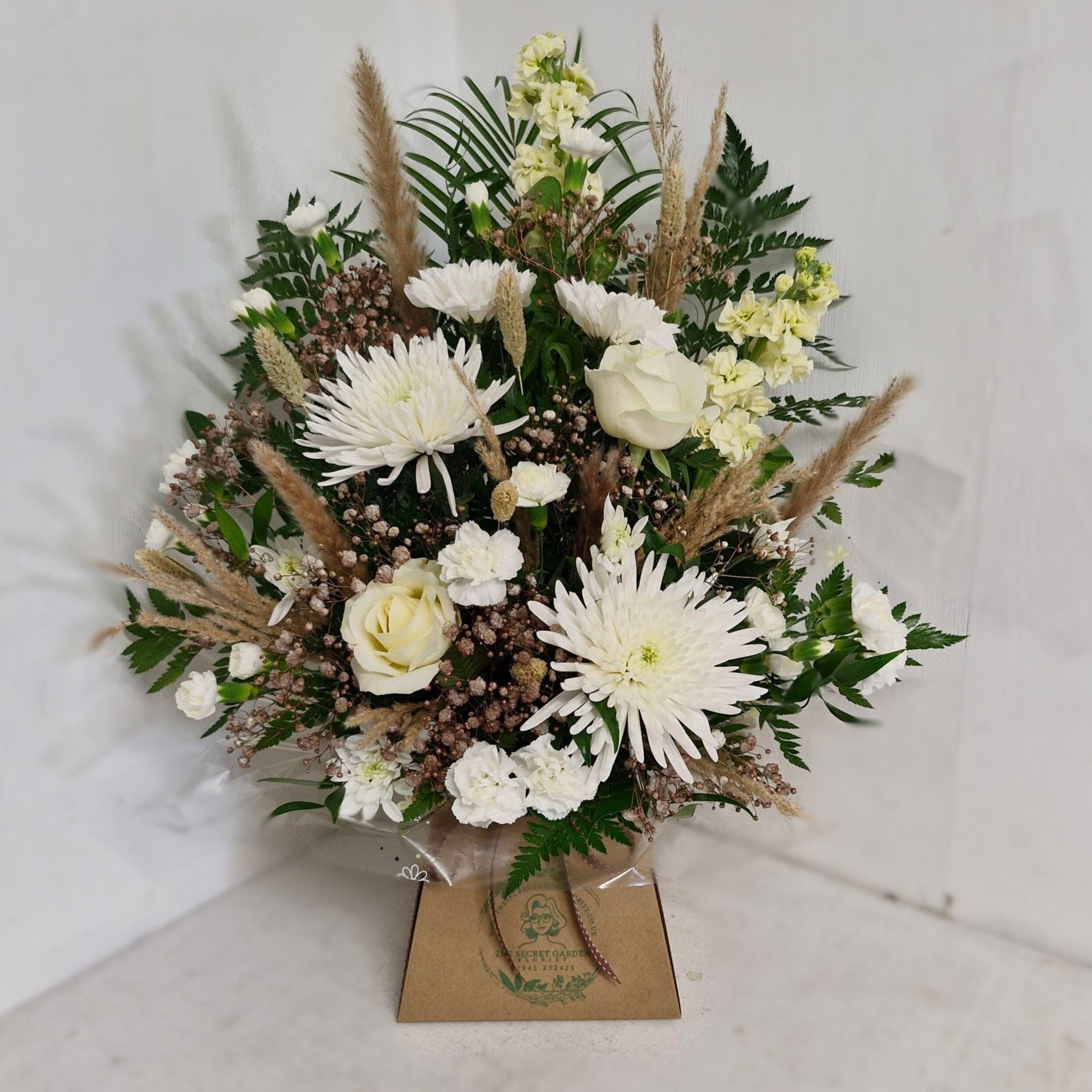 A floral arrangement featuring white flowers such as roses and chrysanthemums, with green foliage and elements of beige and brown, arranged in a Oasis foam base.
