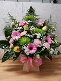 A vibrant floral arrangement featuring pink roses, carnations, and chrysanthemums, with germinis, spray carnations, chrysanthemums, gypsophilia, and mixed greenery, presented in a box with a pink ribbon.