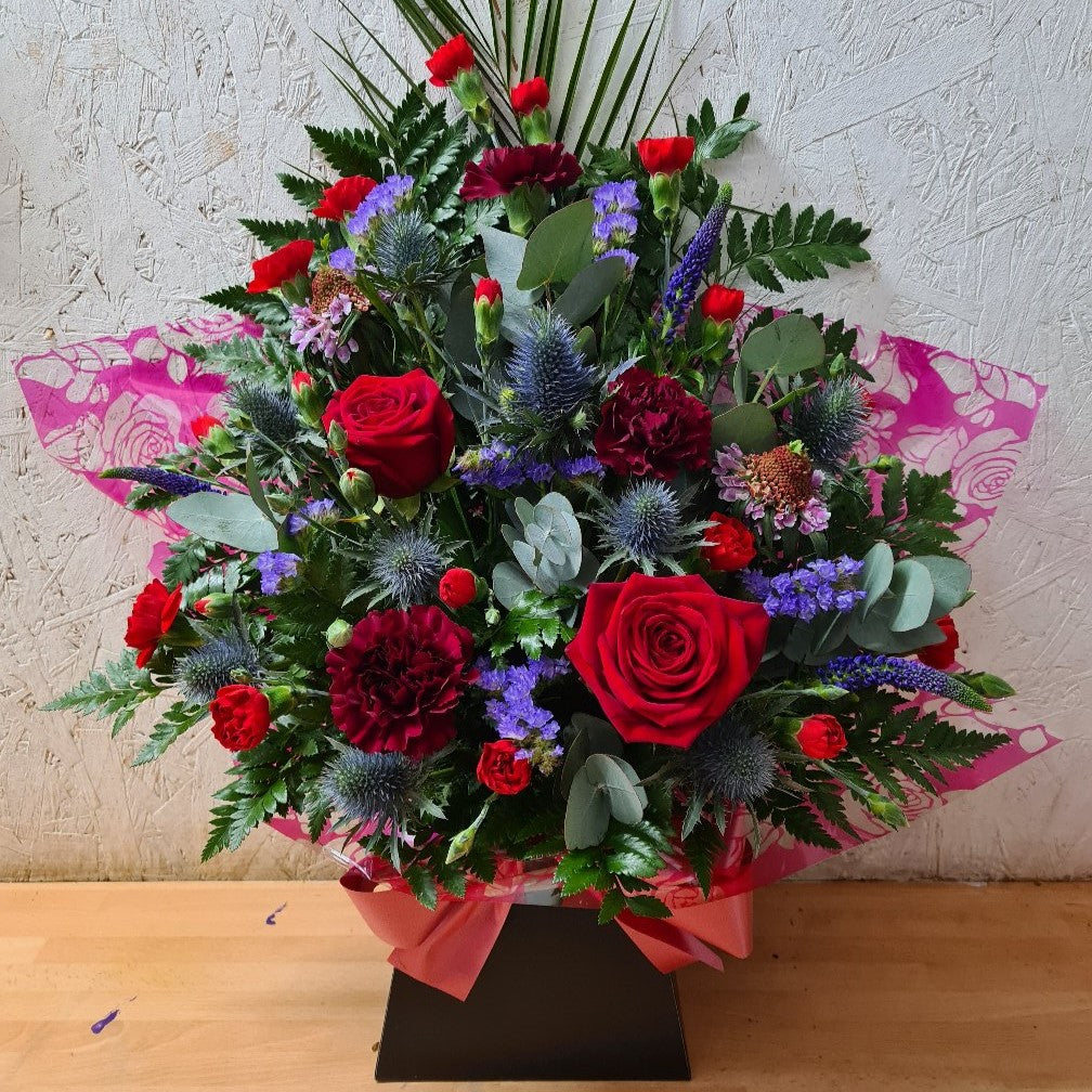 A striking box arrangement of flowers including deep red roses, burgundy flowers, purple accents, and mixed greenery, arranged in oasis floral foam