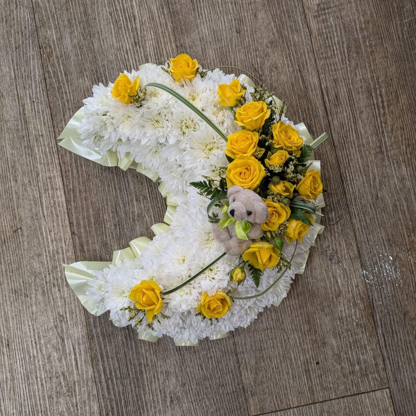 Crescent-shaped floral arrangement with yellow and white flowers and a small teddy bear in the center