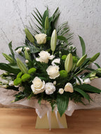 A floral arrangement featuring white roses, lilies, and mixed greenery in a biodegradable floral foam, presented in a box.