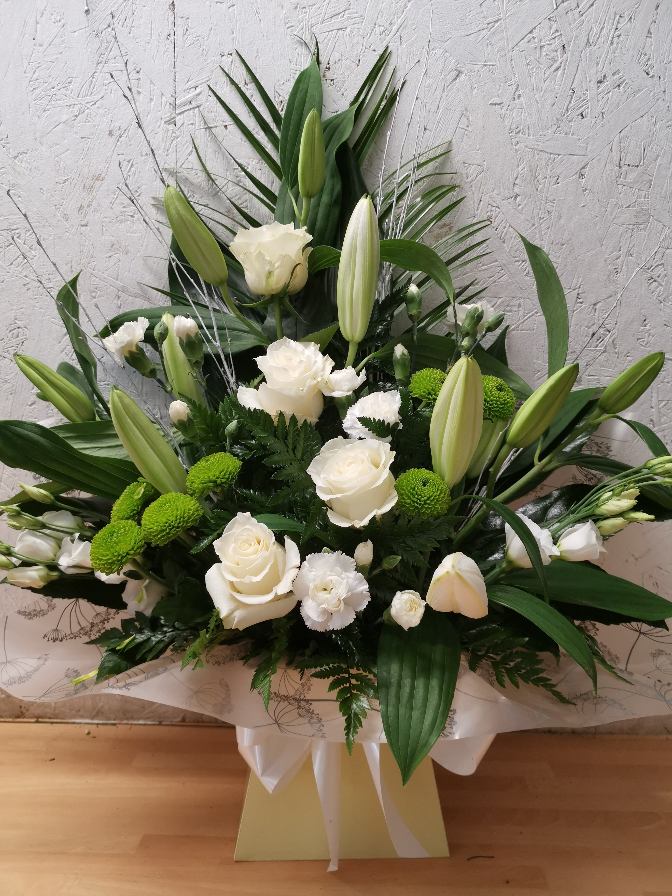 A floral arrangement featuring white roses, lilies, and mixed greenery in a biodegradable floral foam, presented in a box.