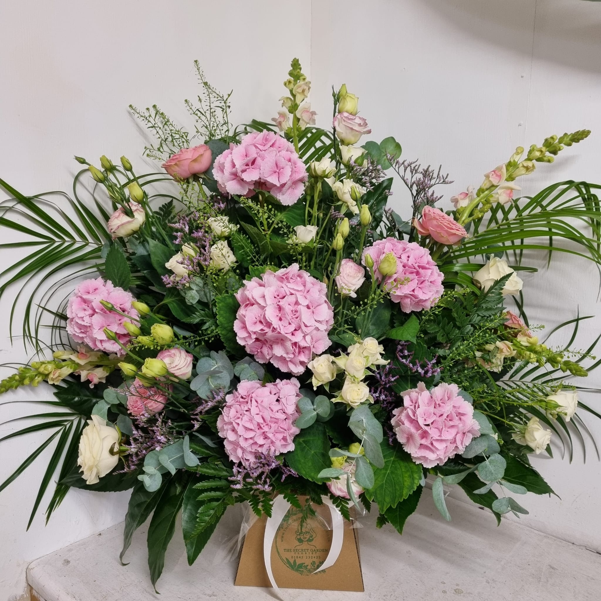 A floral arrangement featuring pale pink and white flowers such as hydrangeas, lisianthus, and roses, with green foliage and spray roses, presented in a floral foam base.