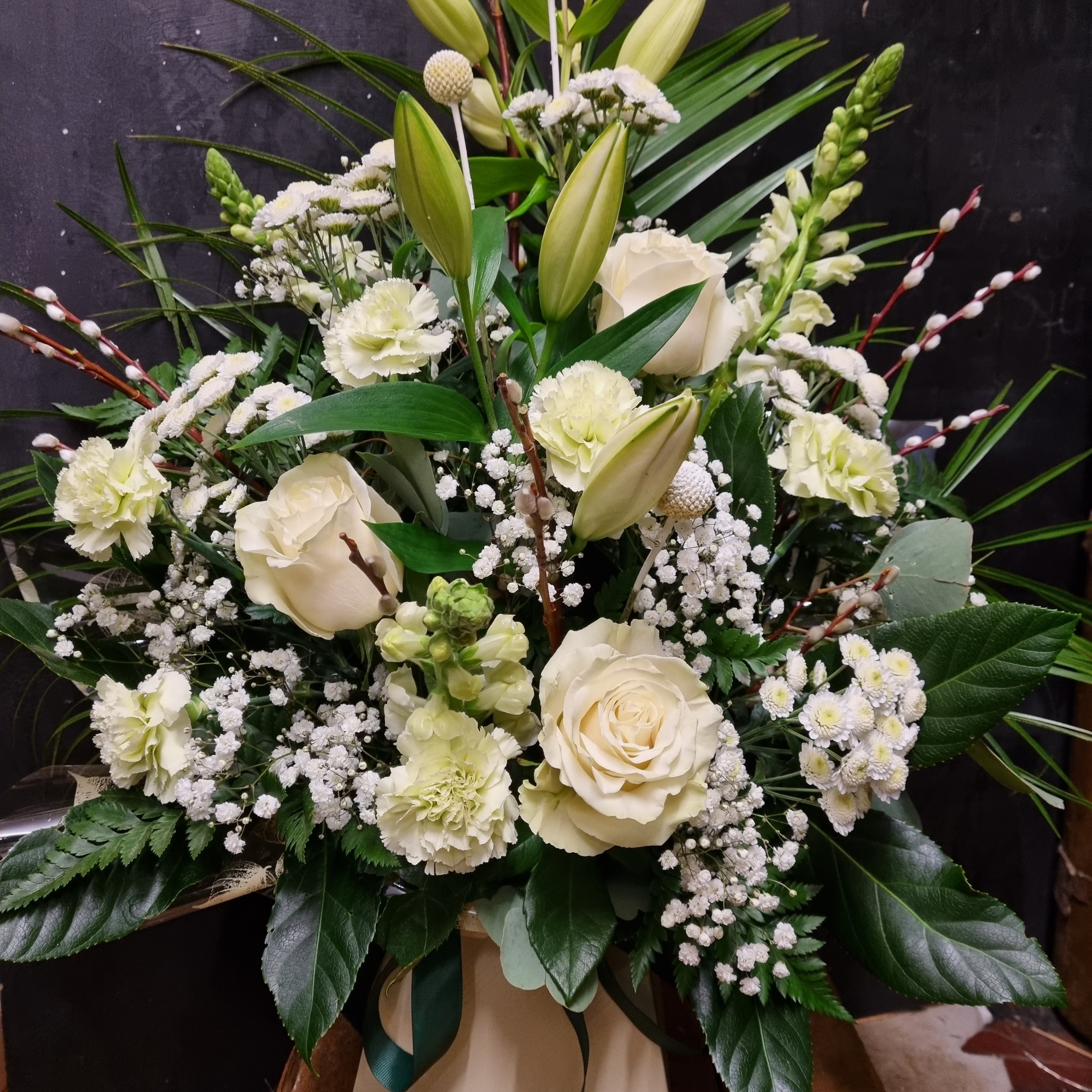 A floral arrangement featuring white roses, lilies, carnations, and other white flowers accompanied by lush green foliage.
