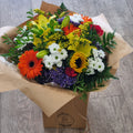 A vibrant handtied bouquet with a variety of flowers including lilies, gerberas, limonium, sunflowers, chrysanthemums, carnations, solidago, and mixed greenery, wrapped in natural brown paper.