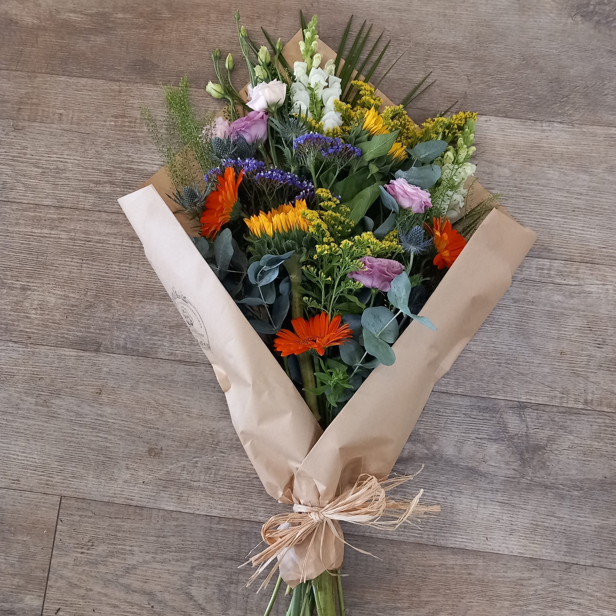 A freshly arranged bouquet of sunflowers, lisianthus, germinis, antirrhinum, solidago, eryngium, limonium, and eucalyptus with other mixed greenery, wrapped in brown paper and tied with a raffia