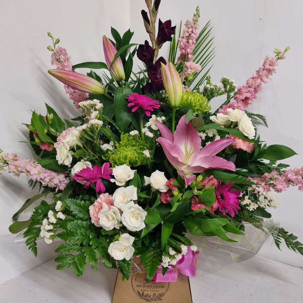 A floral arrangement featuring a variety of flowers including Lilies with other pink, lime, and white flowers, with greenery and arranged in oasis floral foam