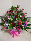 A colorful floral arrangement featuring roses, carnations, germinis, spray carnations, antirrhinum, statice, freesia, gypsophilia, and mixed greenery, arranged in oasis floral foam