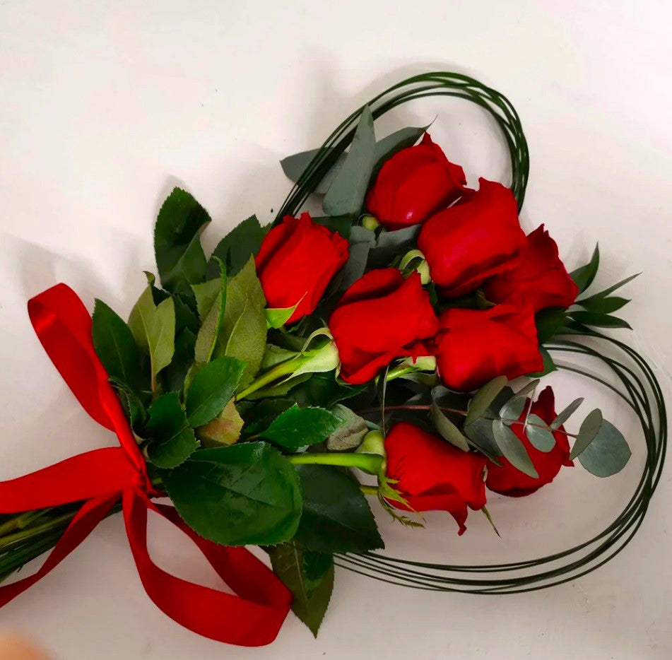 A bouquet of large red roses arranged to form a heart shape with steel grass and mixed greenery, tied with a red ribbon.