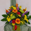 A vibrant floral arrangement featuring a variety of flowers such as Chrysanthemums, Gerberas, Carnations, and other greenery, arranged in watered floral foam.