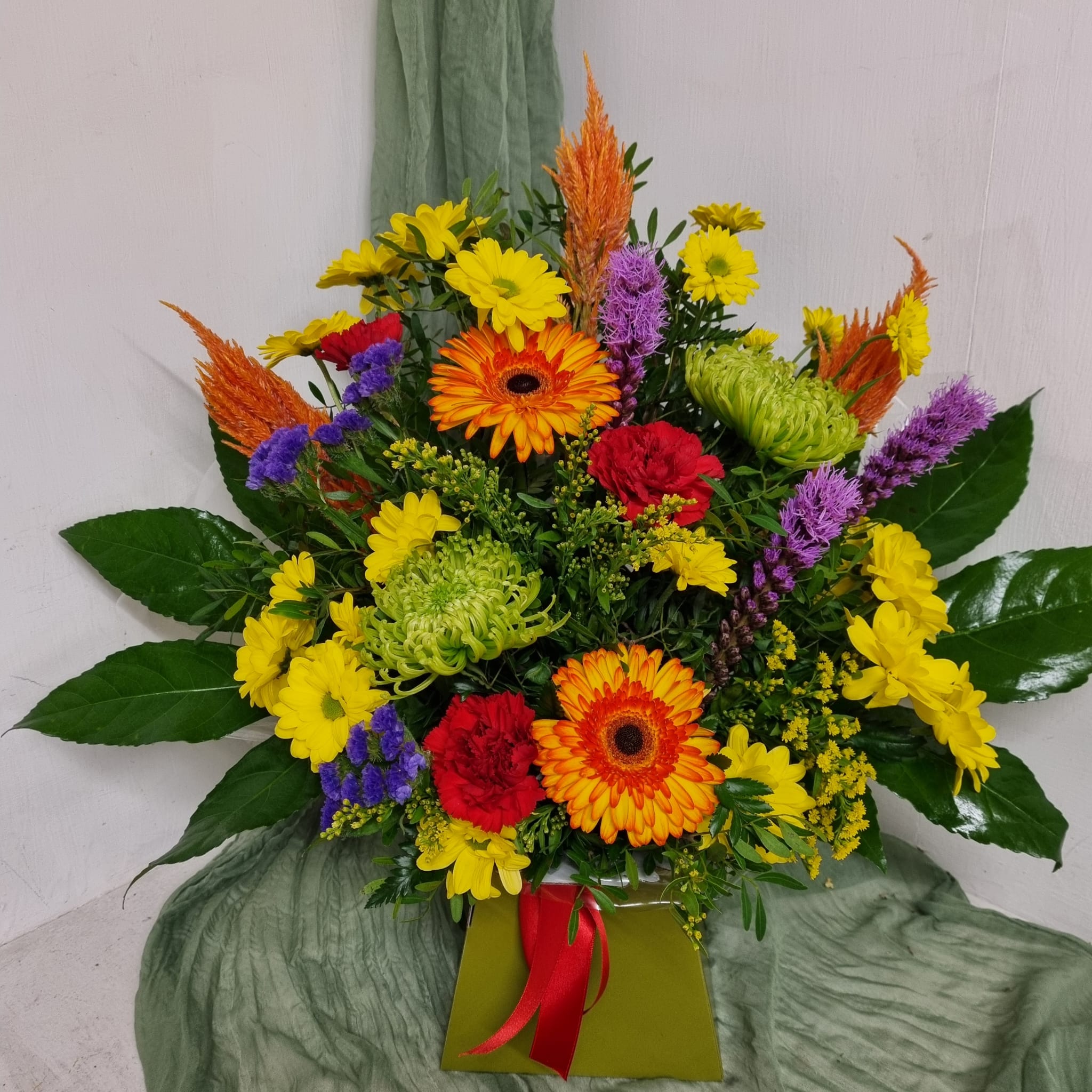 A vibrant floral arrangement featuring a variety of flowers such as Chrysanthemums, Gerberas, Carnations, and other greenery, arranged in watered floral foam.