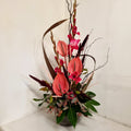A floral arrangement featuring pink anthuriums, green foliage, and decorative elements in a tall display vase.