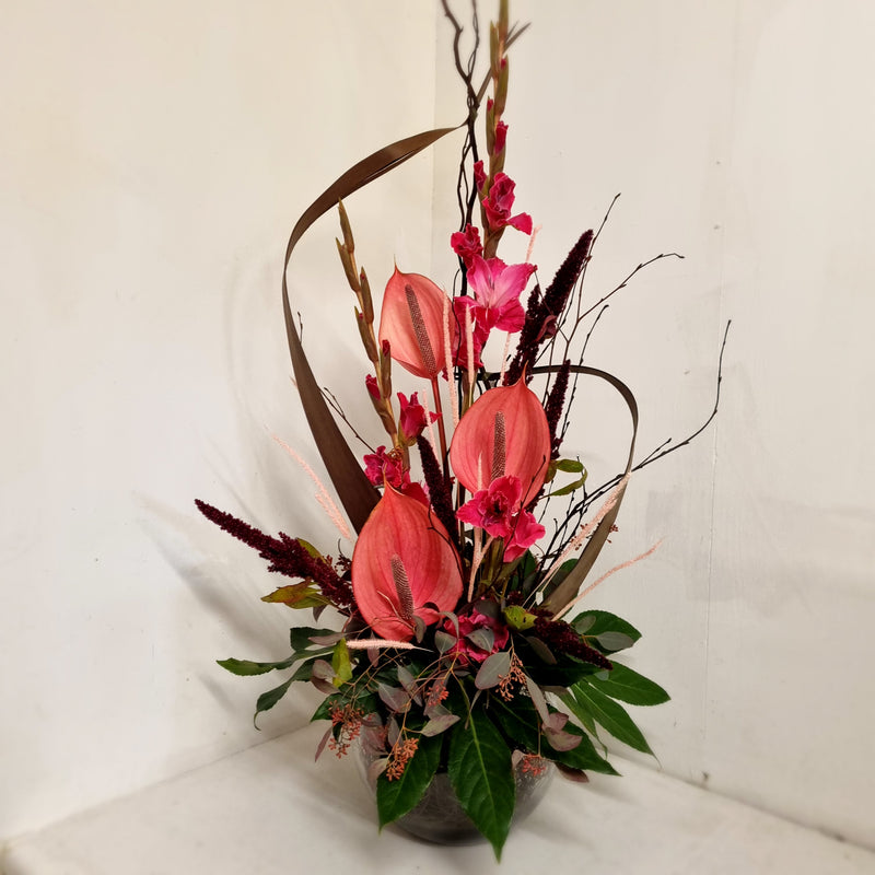 A floral arrangement featuring pink anthuriums, green foliage, and decorative elements in a tall display vase.