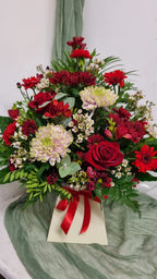 A floral arrangement featuring roses, chrysanthemums, and other mixed flowers in shades of red and pink, with greenery arranged in oasis floral foam