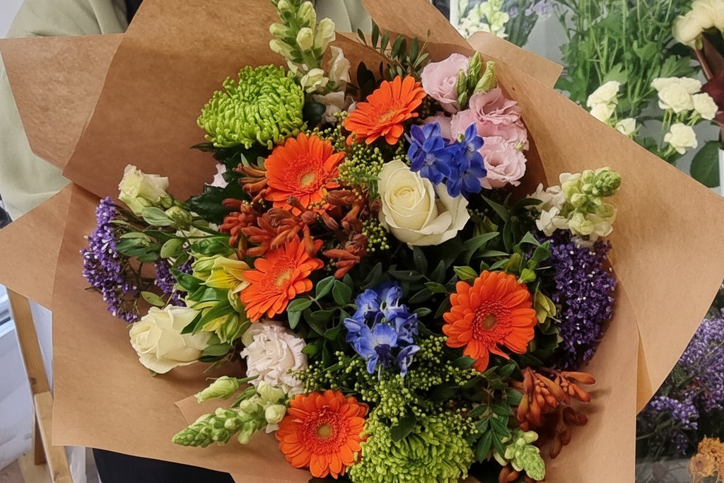 A hand-tied bouquet with a variety of colorful flowers including oranges, purples, and limes, wrapped in brown paper.