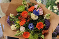 A hand-tied bouquet with a variety of colorful flowers including oranges, purples, and limes, wrapped in brown paper.