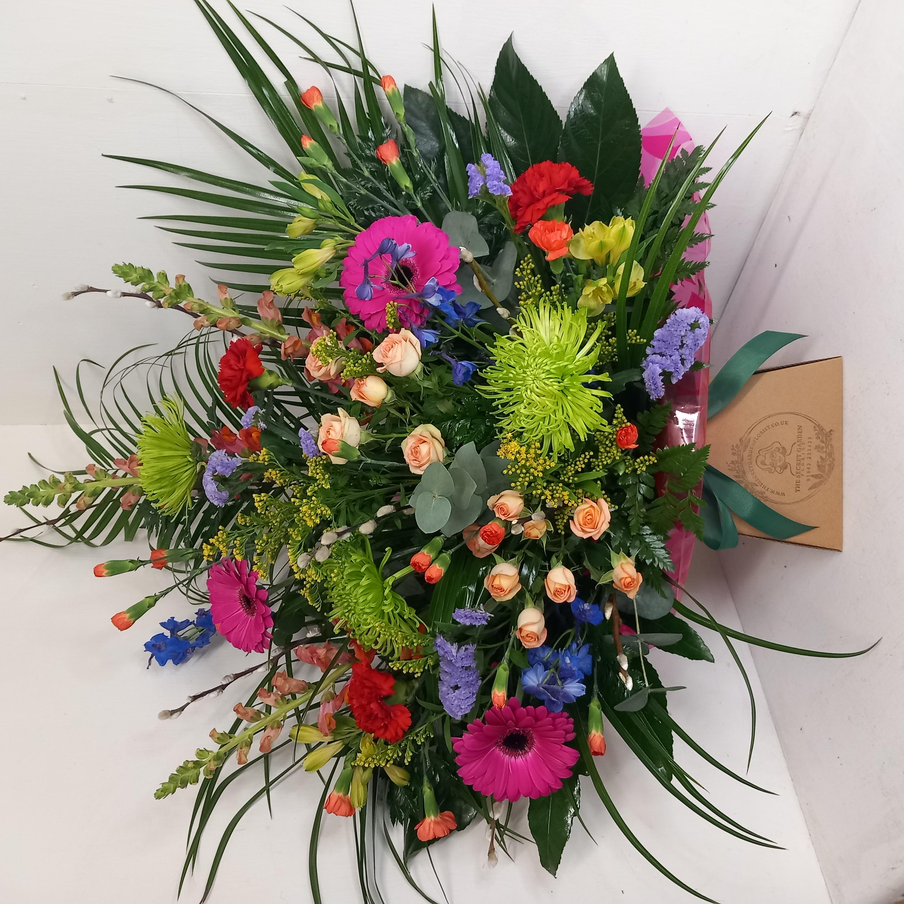 A vibrant floral arrangement featuring a variety of flowers such as spray roses, Germinis, Chrysanthemum blooms, Alstromeria, Delphinium, Carnations, Spray carnations, Antirrhinums, Statice, Solidago, and mixed greenery, presented in a box.