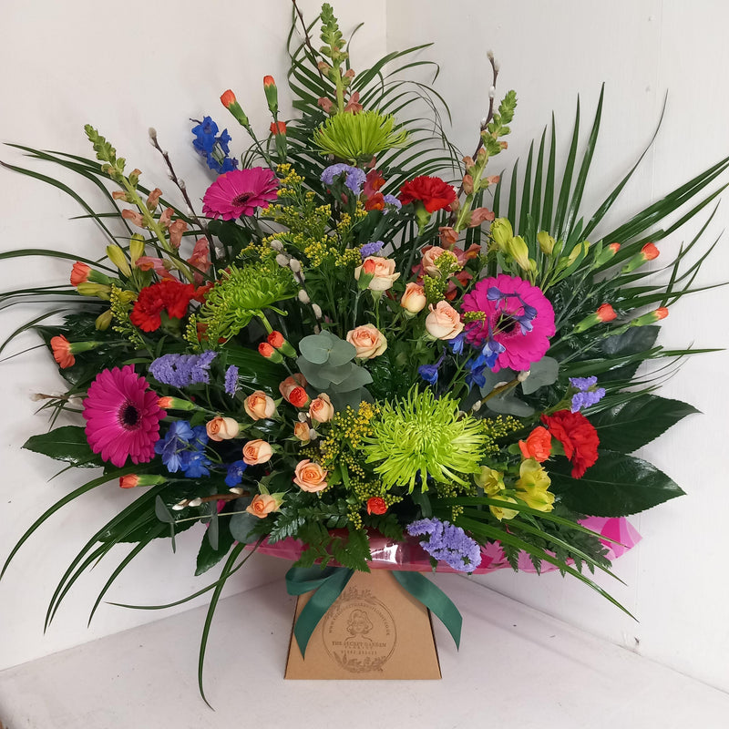 A vibrant floral arrangement featuring a variety of flowers such as spray roses, Germinis, Chrysanthemum blooms, Alstromeria, Delphinium, Carnations, Spray carnations, Antirrhinums, Statice, Solidago, and mixed greenery, presented in a box.