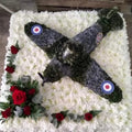 A 2ft by 2ft design board featuring a 3D model of a Spitfire plane with green and grey colours, surrounded by white flowers and a small spray of red roses.