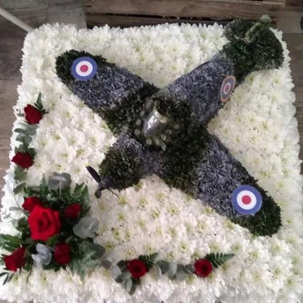 A 2ft by 2ft design board featuring a 3D model of a Spitfire plane with green and grey colours, surrounded by white flowers and a small spray of red roses.