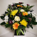 A vibrant floral arrangement featuring bright yellow and purple flowers, such as Chrysanthemums and Roses, with mixed greenery in a box.