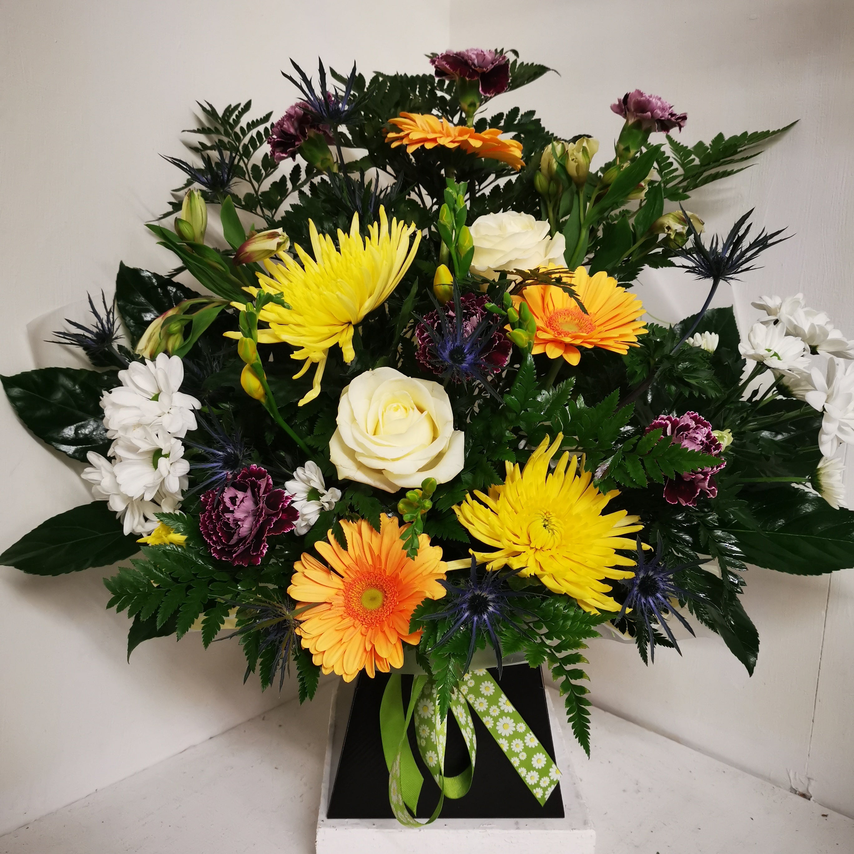A vibrant floral arrangement featuring bright yellow and purple flowers, such as Chrysanthemums and Roses, with mixed greenery in a box.