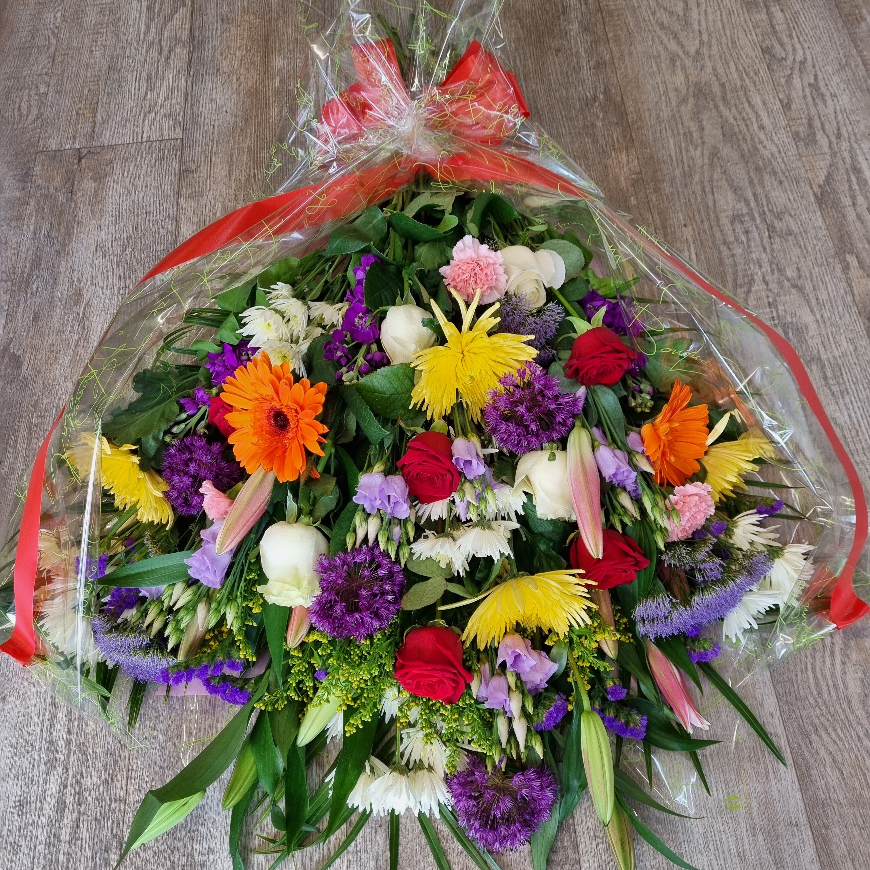 A vibrant bouquet of assorted flowers including roses, chrysanthemums, gerberas, carnations, and other greenery, wrapped in cello and tied with ribbons.