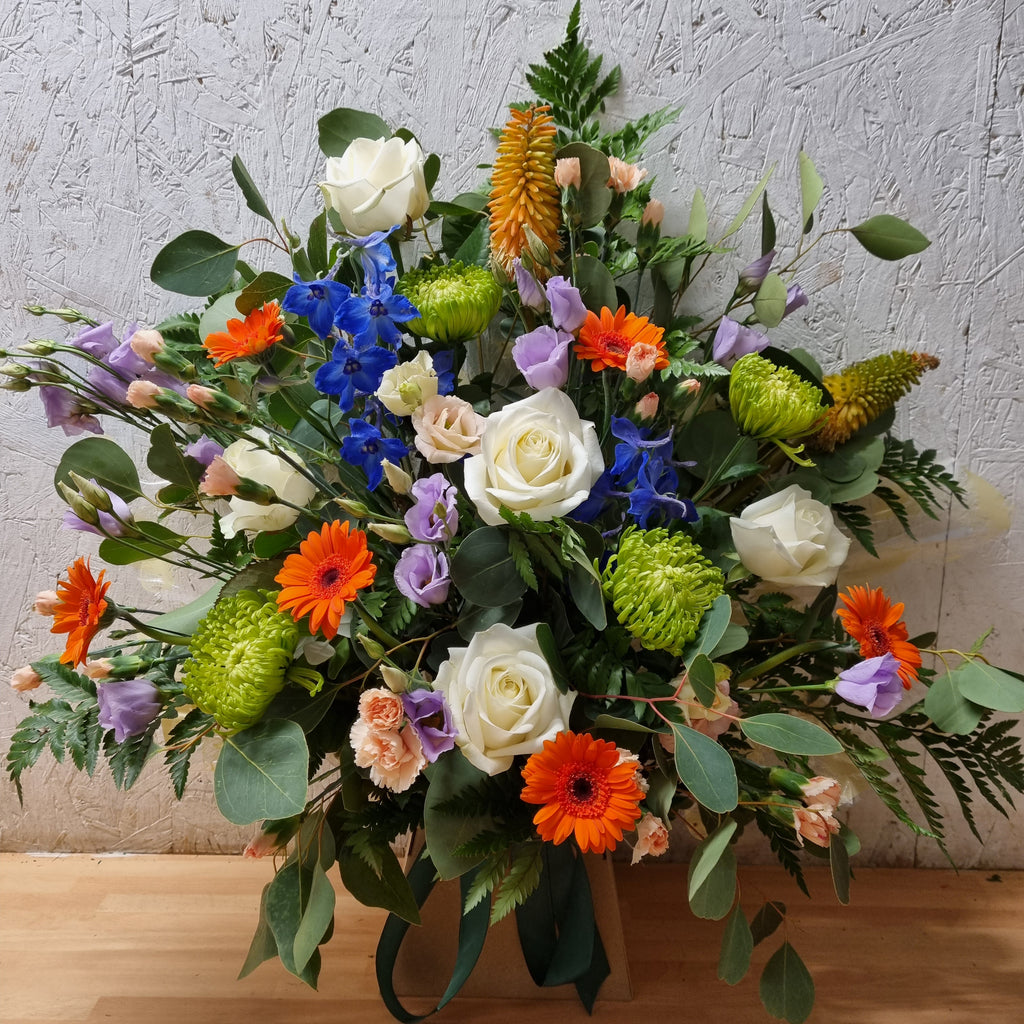 A vibrant floral arrangement featuring a variety of flowers including roses, chrysanthemums, germinis, lisianthus, spray carnations, delphinium, kniphofia, and mixed greenery, displayed in eco-friendly floral foam.