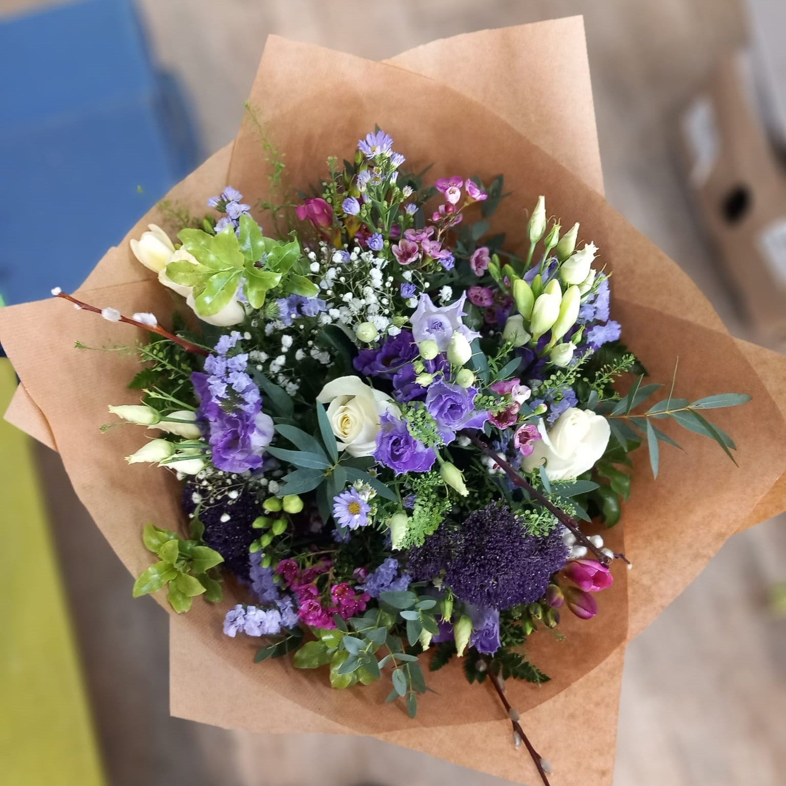 A hand-tied bouquet of purple and white flowers with a mix of greenery, wrapped in natural paper.