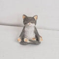A small gray and white cat figurine sitting cross-legged in a meditative pose, photographed against a plain light gray background.