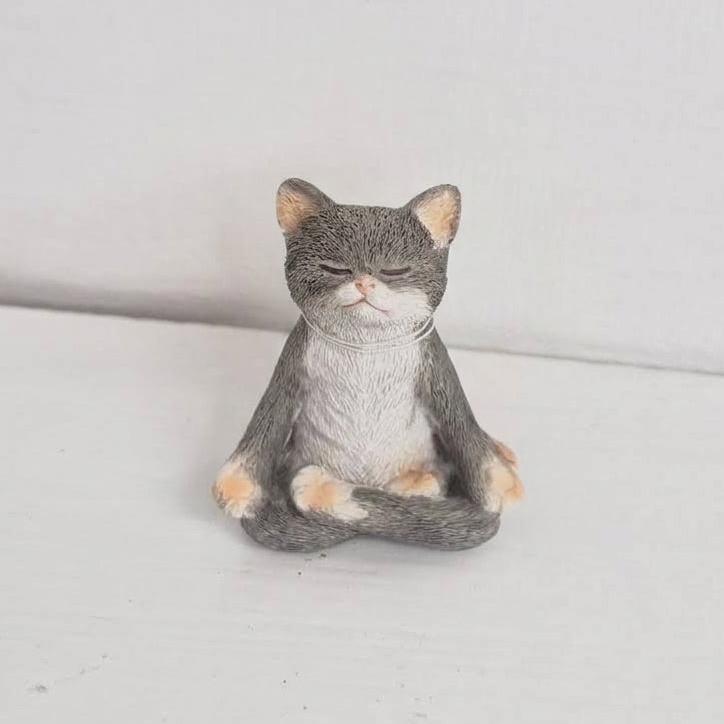 A small gray and white cat figurine sitting cross-legged in a meditative pose, photographed against a plain light gray background.