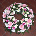 A round pink and white floral wreath composed of various flowers including roses and gerberas, with green leaves