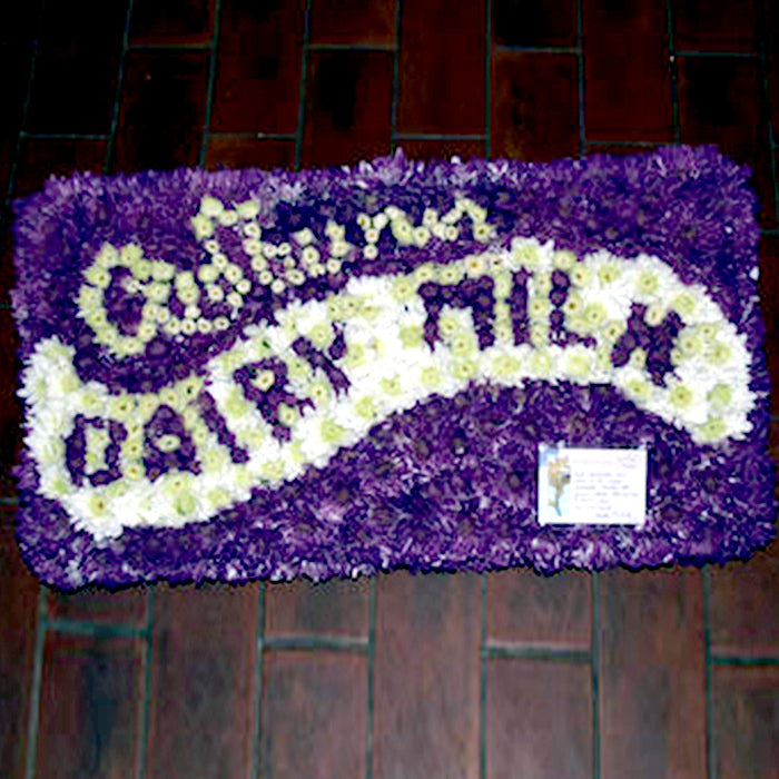 A floral arrangement designed to resemble a carton of Dairy Milk, predominantly purple and white in color, displayed on a flat surface.