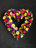 A brightly colored floral arrangement in the shape of an open heart, featuring a mix of flowers including yellow, purple, and pink.
