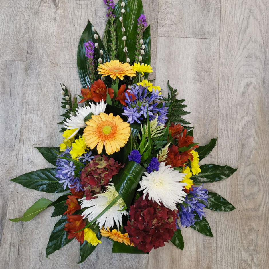 A floral tribute in a teardrop shape, featuring a variety of flowers, including yellow, orange, and blue, complemented by green foliage.