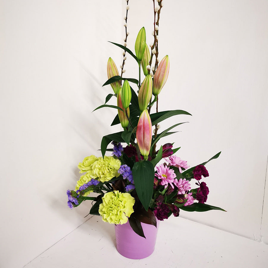 A tall slim floral arrangement with a variety of flowers including lilies, carnations, and other foliage, arranged in a ceramic container.