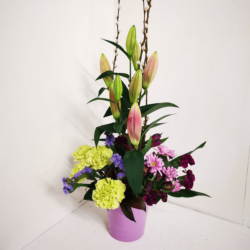 A tall slim floral arrangement with a variety of flowers including lilies, carnations, and other foliage, arranged in a ceramic container.